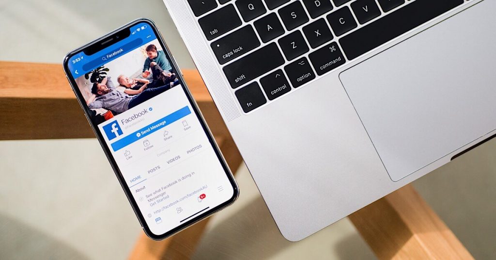An iPhone X with the Facebook app on-screen and a laptop to the right.