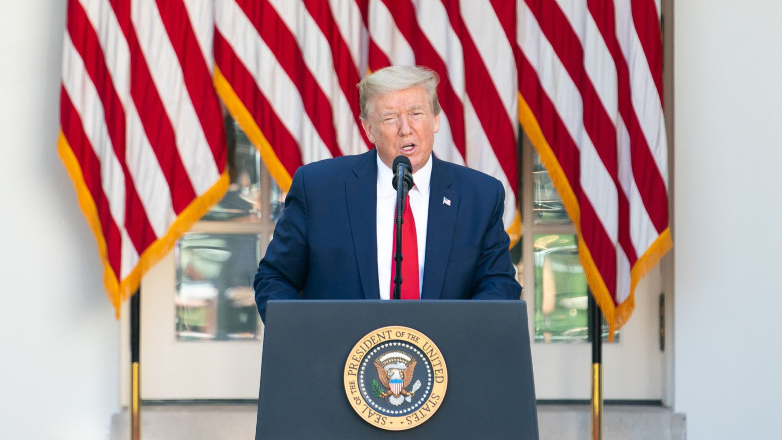 Trump with light-colored hair in a navy suit and red tie speaks at a podium bearing the Presidential Seal, with multiple American flags draped behind him.