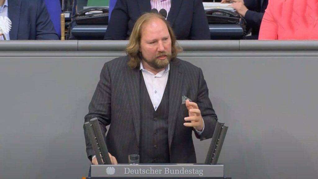 Hofreiter with long hair and a beard is speaking at a podium labeled "Deutscher Bundestag." He is wearing a pinstriped suit.