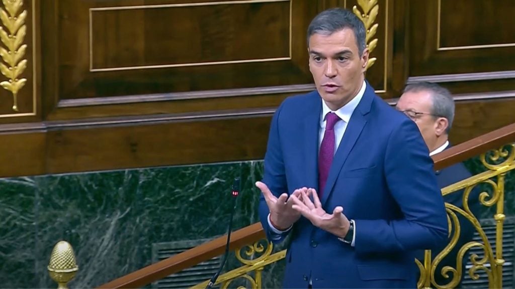 Pedro Sanchez in a red tie speaking at a podium, with ornate wooden and gold decor in the background.