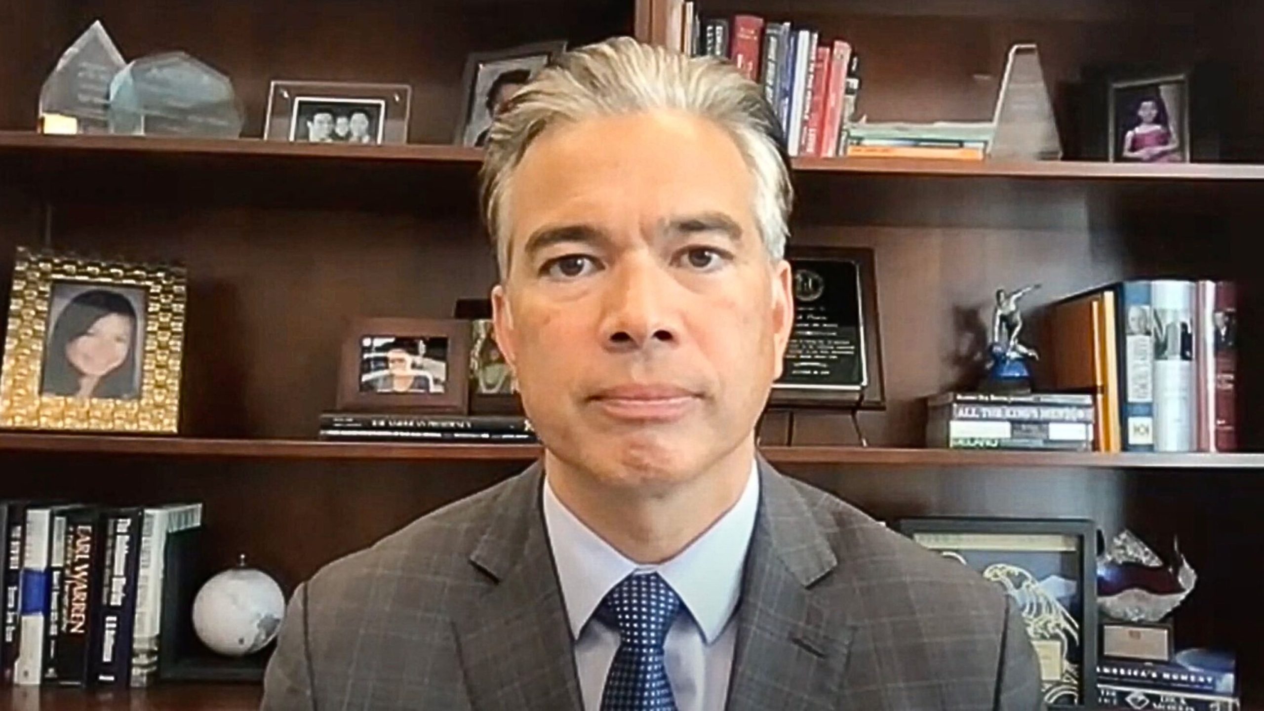 California AG Rob Bonta in a suit and tie sitting in front of a wooden bookshelf filled with books, framed photos, and various decorative items.