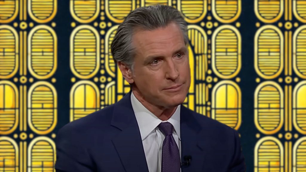 Gavin Newsom in a suit and tie with a serious expression, in front of a patterned background with yellow and black geometric shapes.