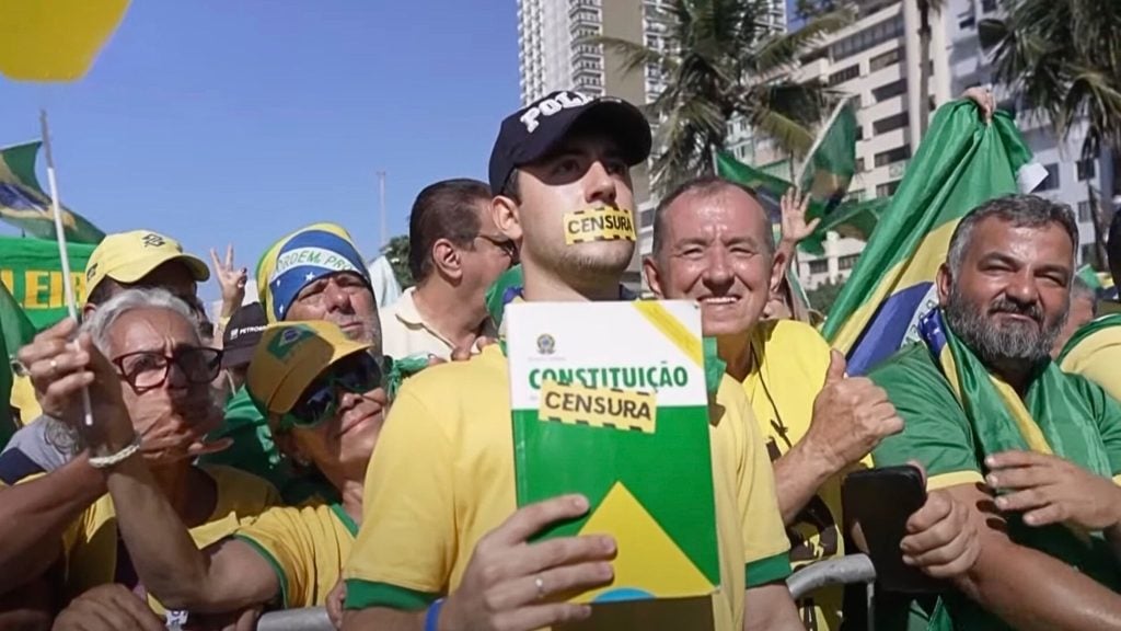 Crowd of people wearing yellow and green, holding a book labeled "Constituição" with a "Censura" strip, and a man with a "Censura" strip over his mouth.