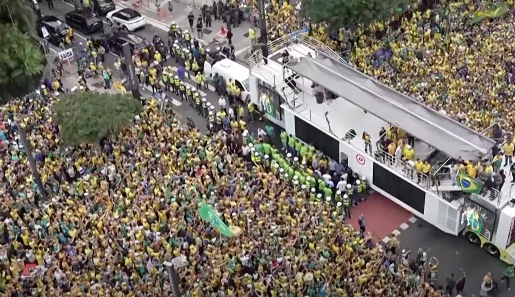 A large crowd gathers around a vehicle covered in banners and flags, with many people dressed in yellow and green, including law enforcement personnel forming a line.