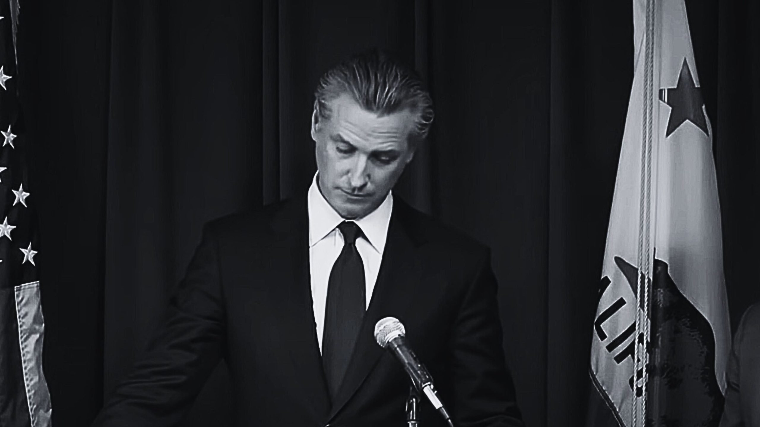 Governor Gavin Newsom in a suit standing at a podium with microphones, between the United States and California flags.