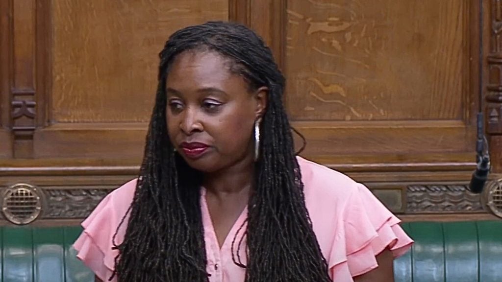 Dawn Butler with long braided hair and pink blouse standing in a wooden-paneled room.