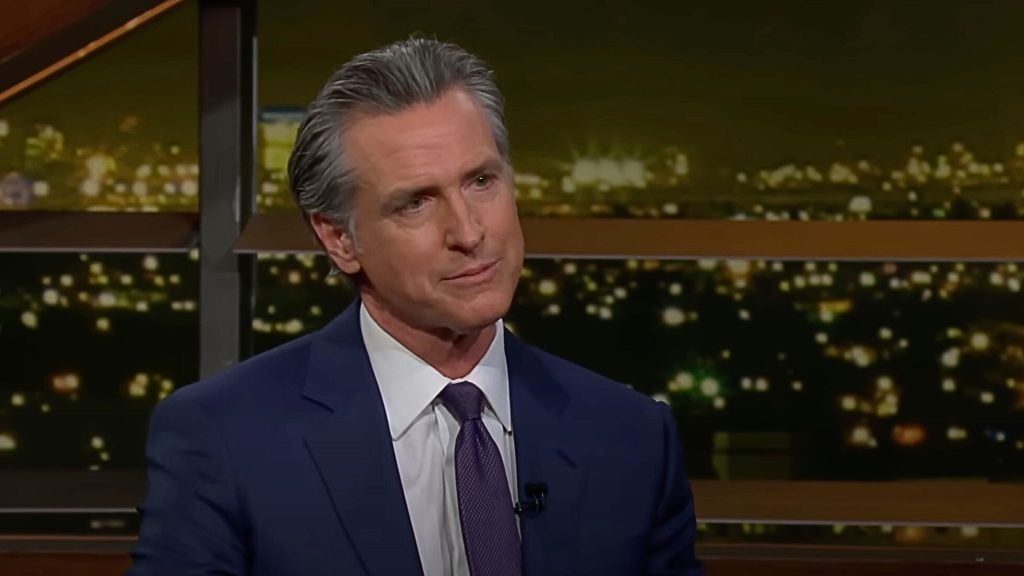 Gavin Newsom in a suit and tie sitting indoors with a cityscape at night in the background.