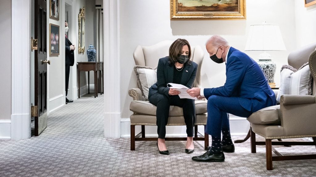 President Biden and Vice President Harris in formal attire sit on armchairs, both wearing masks, reviewing documents. The setting is a well-lit room with framed artwork and a lamp in the background. Another person stands in the hallway.