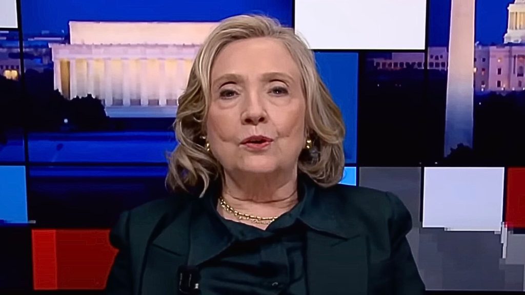 Hillary Clinton with blond hair speaking on TV, with a backdrop of the Lincoln Memorial and Washington Monument at night.