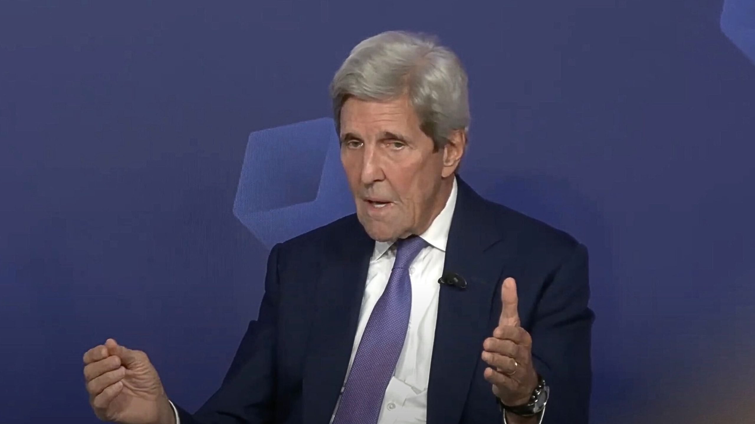 John Kerry in a suit and tie speaking with hand gestures against a dark blue background.