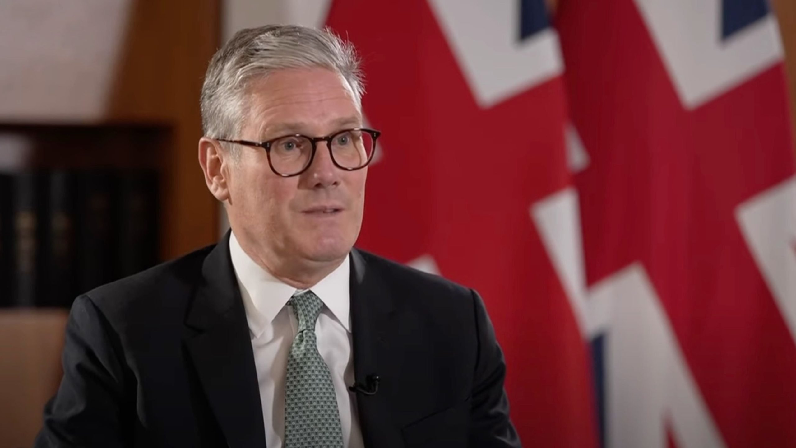 Keir Starmer in suit and tie with glasses sits in front of a blurred background with partial view of two British flags.