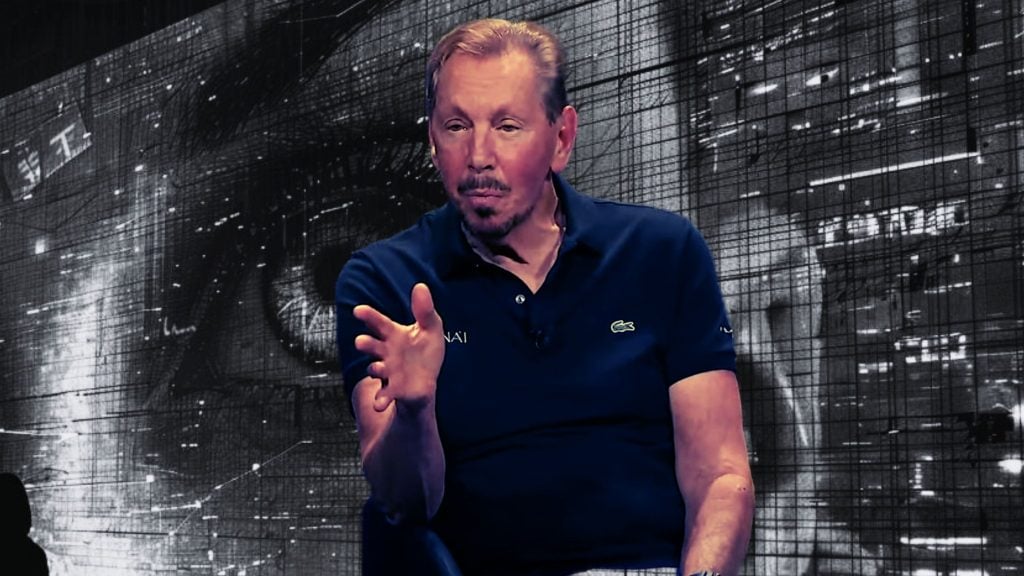 Larry Ellison blue polo shirt gestures while speaking, with a large graphic of an eye and digital elements in the background.