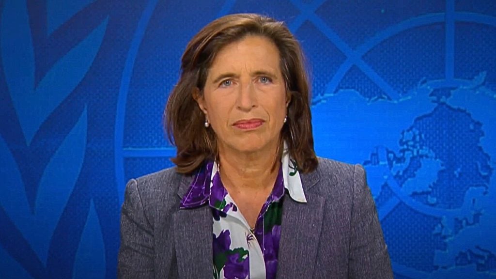 Melissa Fleming with shoulder-length brown hair in a gray blazer and floral blouse, in front of a blue background with a global map design.