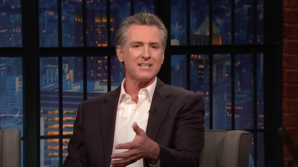 Gavin Newsom talking on a late-night talk show set, with a cityscape visible through large windows in the background.