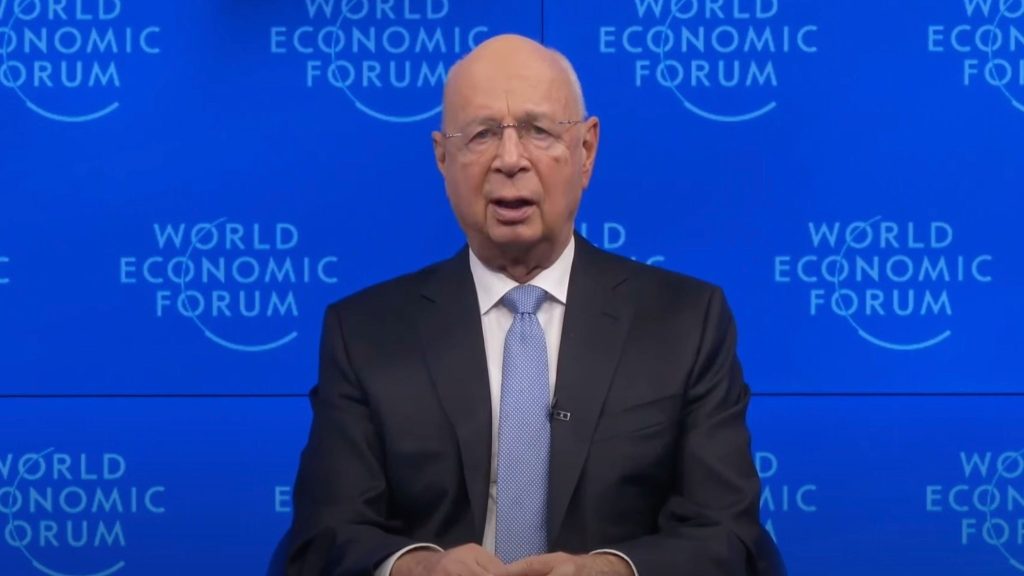 Schwab in a suit and tie speaking in front of a backdrop with the "World Economic Forum" logo.