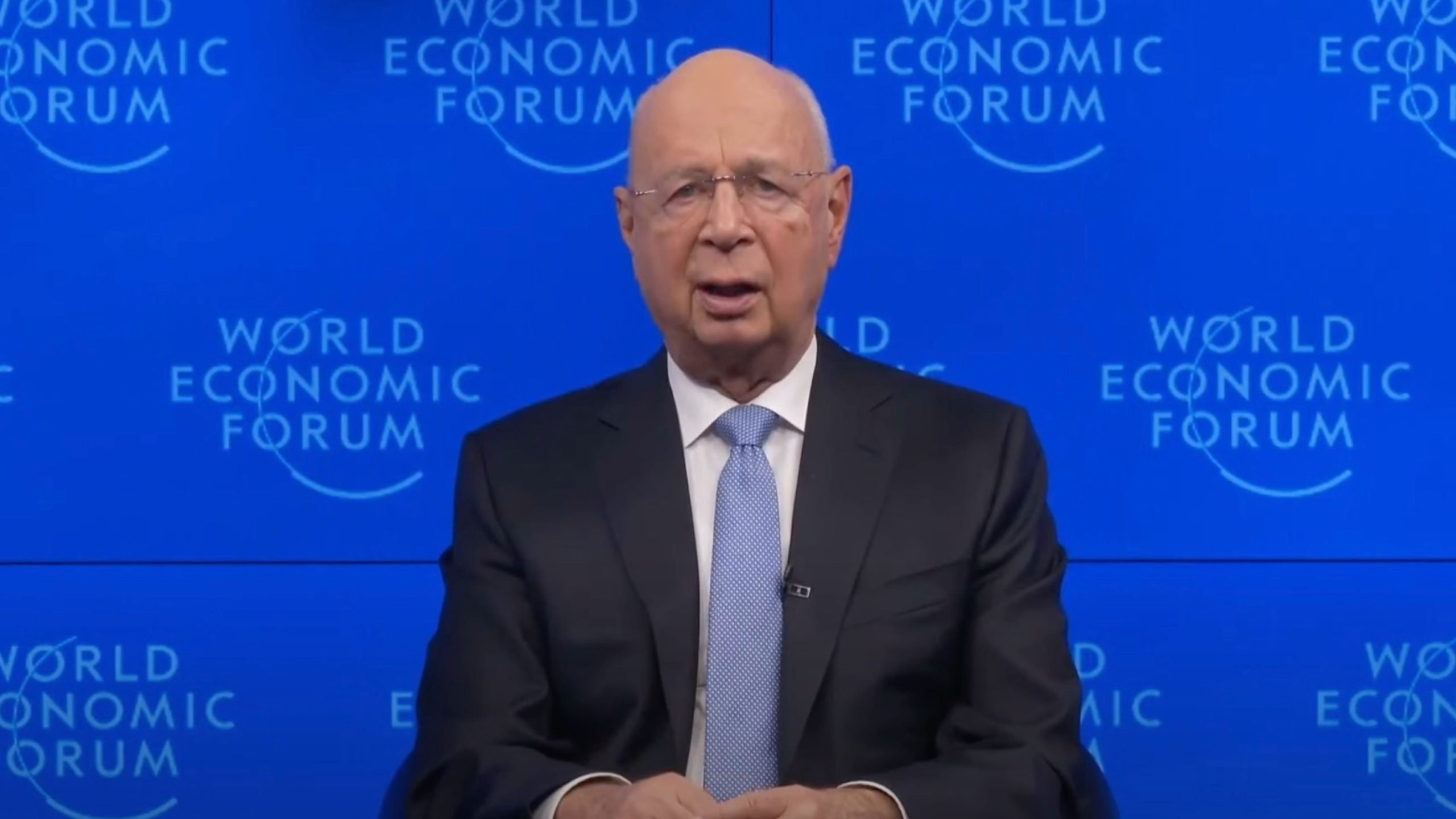 Schwab in a suit and tie speaking in front of a backdrop with the "World Economic Forum" logo.