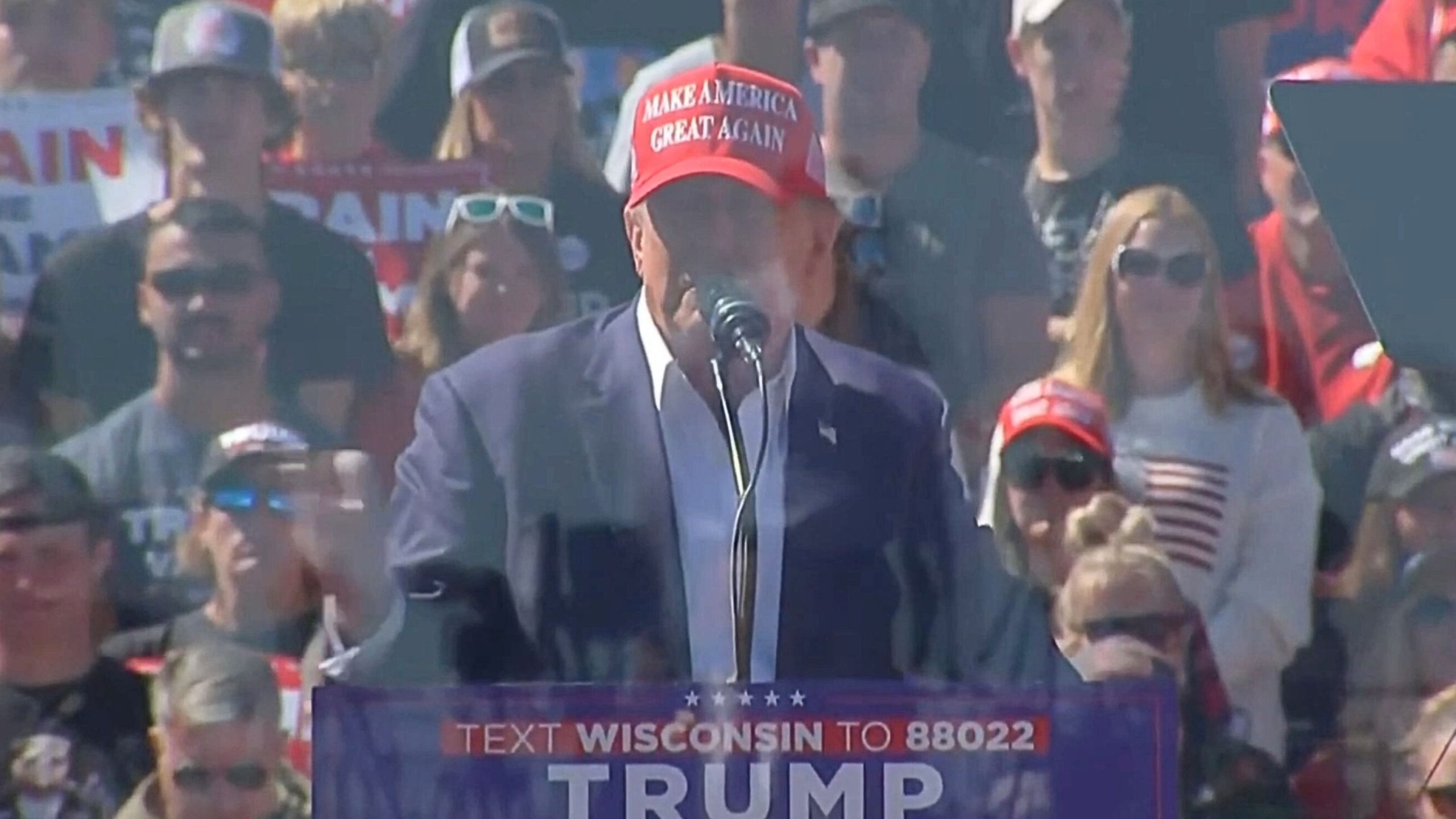 President Trump wearing a "Make America Great Again" hat speaks at a rally, with a crowd of supporters in the background.