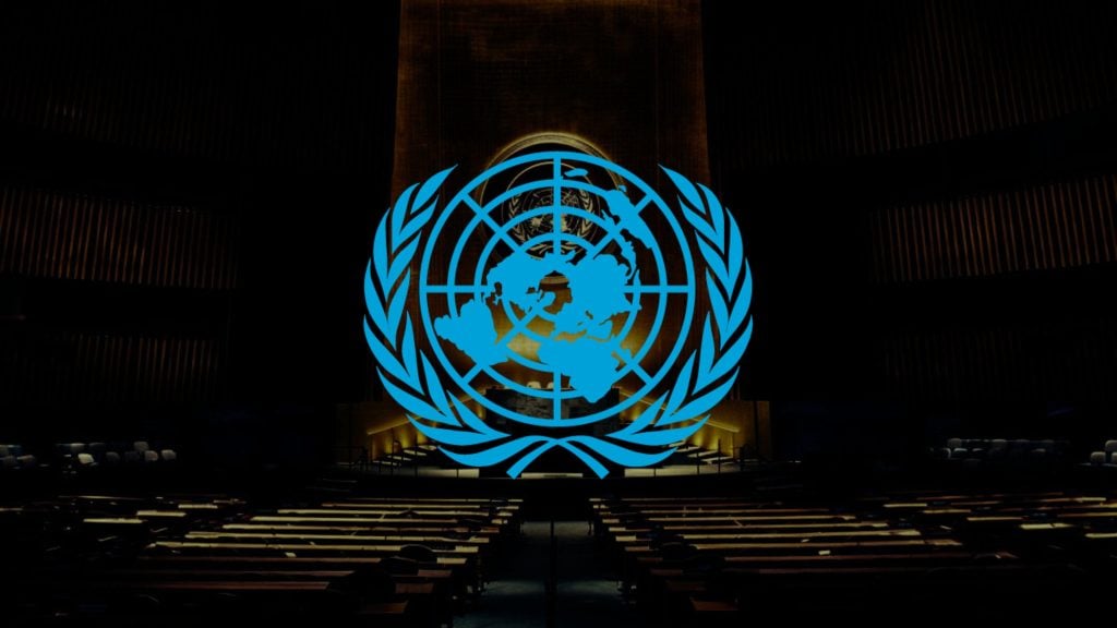 Blue United Nations logo overlaid on a dimly lit conference hall with rows of seating.