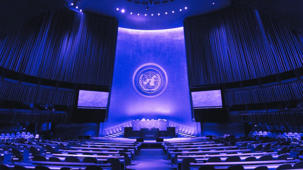 A large, dimly lit conference hall with rows of desks and seats, featuring a central podium and a large emblem on the wall illuminated in blue light.