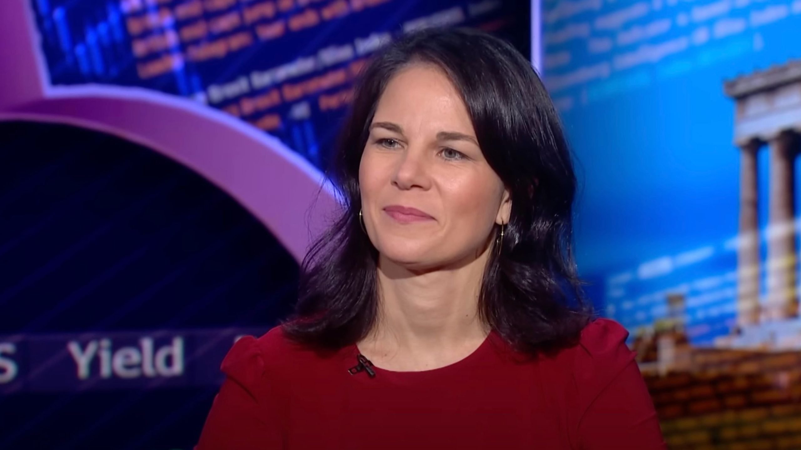 Annalena Baerbock with dark hair and a red top, with blurred text and architectural elements in the background.