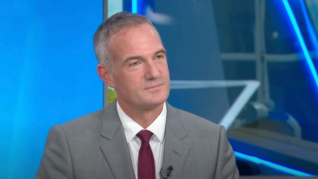 Peter Kyle in a gray suit with a red tie sitting in a room with blue background lighting.