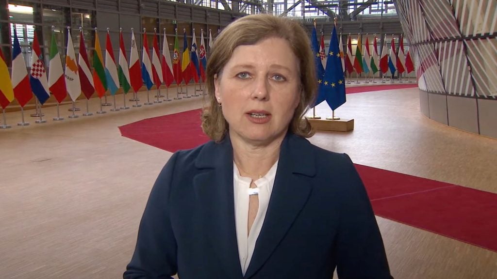 Vera Jourova with light brown hair is speaking in an indoor setting with numerous national flags and a red carpet in the background.