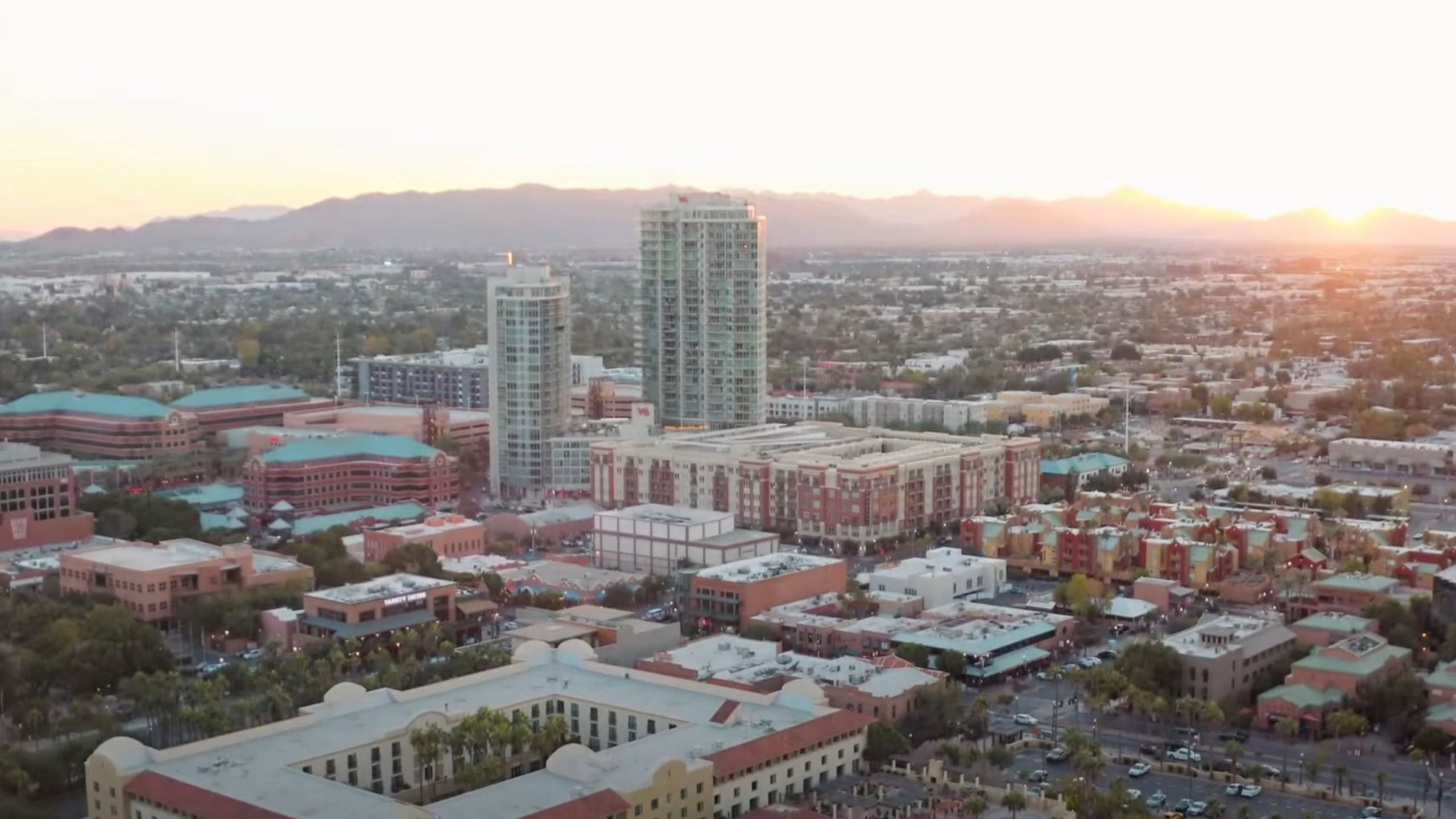 Aerial view of an Arizona cityscape at sunset, featuring tall buildings, low-rise structures, and distant mountains.