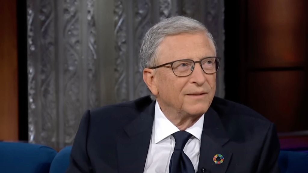 Bill Gates wearing glasses, a suit, and a Global Goals pin on his lapel, sitting against a decorative background.