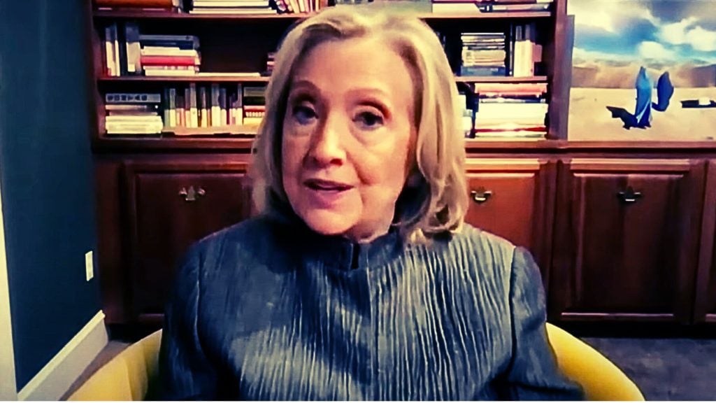 Hillary Clinton in front of a bookshelf, speaking, with a painting on the right side.