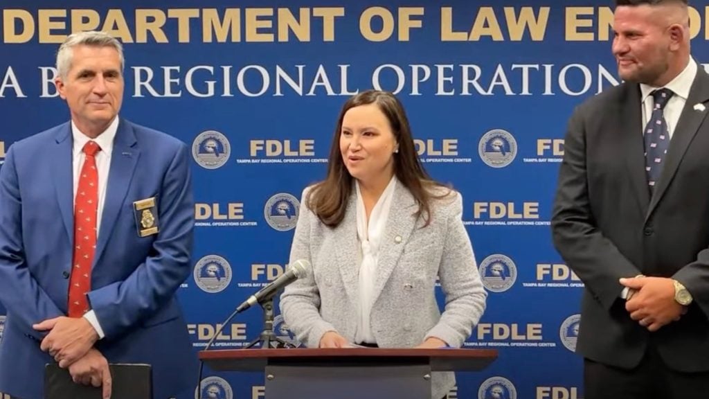 Ashley Moody standing at a podium with a blue backdrop reading "DEPARTMENT OF LAW ENFORCEMENT FDLE TAMPA BAY REGIONAL OPERATIONS CENTER."