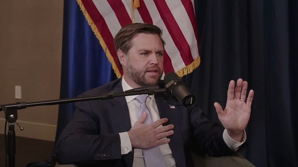 JD Vance speaking into a microphone with an American flag in the background.