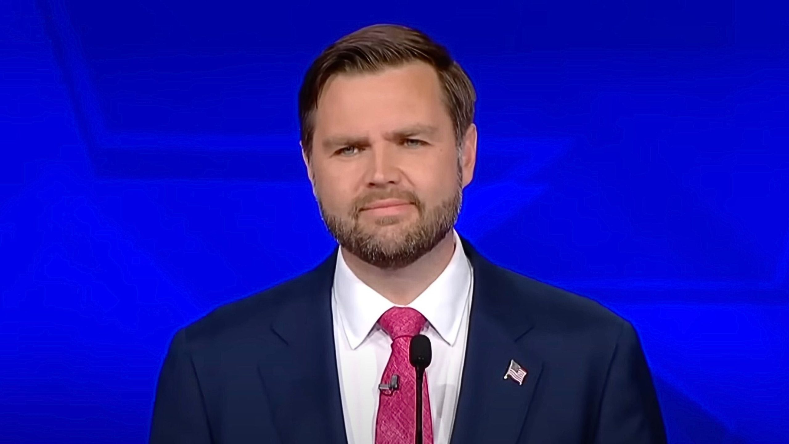 J.D Vance in a suit with a pink tie, standing at a podium against a blue background.