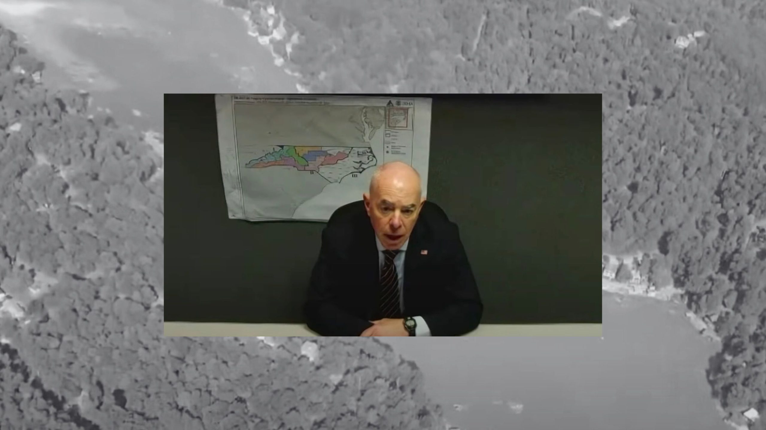 Mayorkas sitting at a table with a map on the wall behind him, superimposed on a grayscale aerial image of a forested landscape.