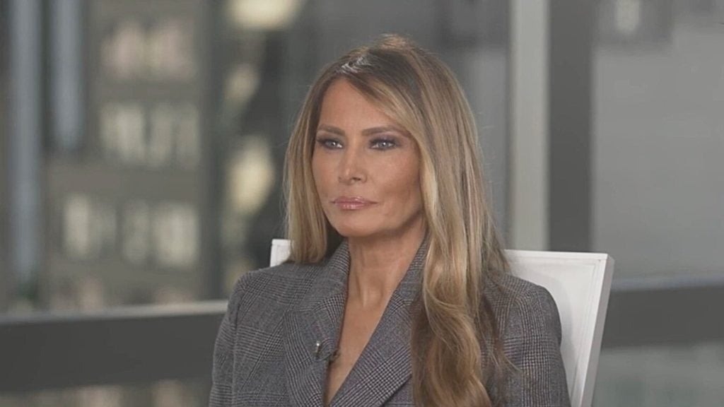 Melania Trump wearing a gray plaid blazer, sitting indoors.