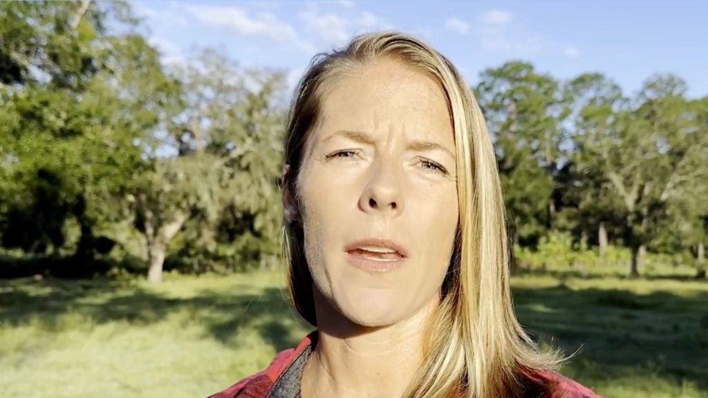 Morrow with long blonde hair standing outdoors in a grassy area with trees in the background, under a clear blue sky.