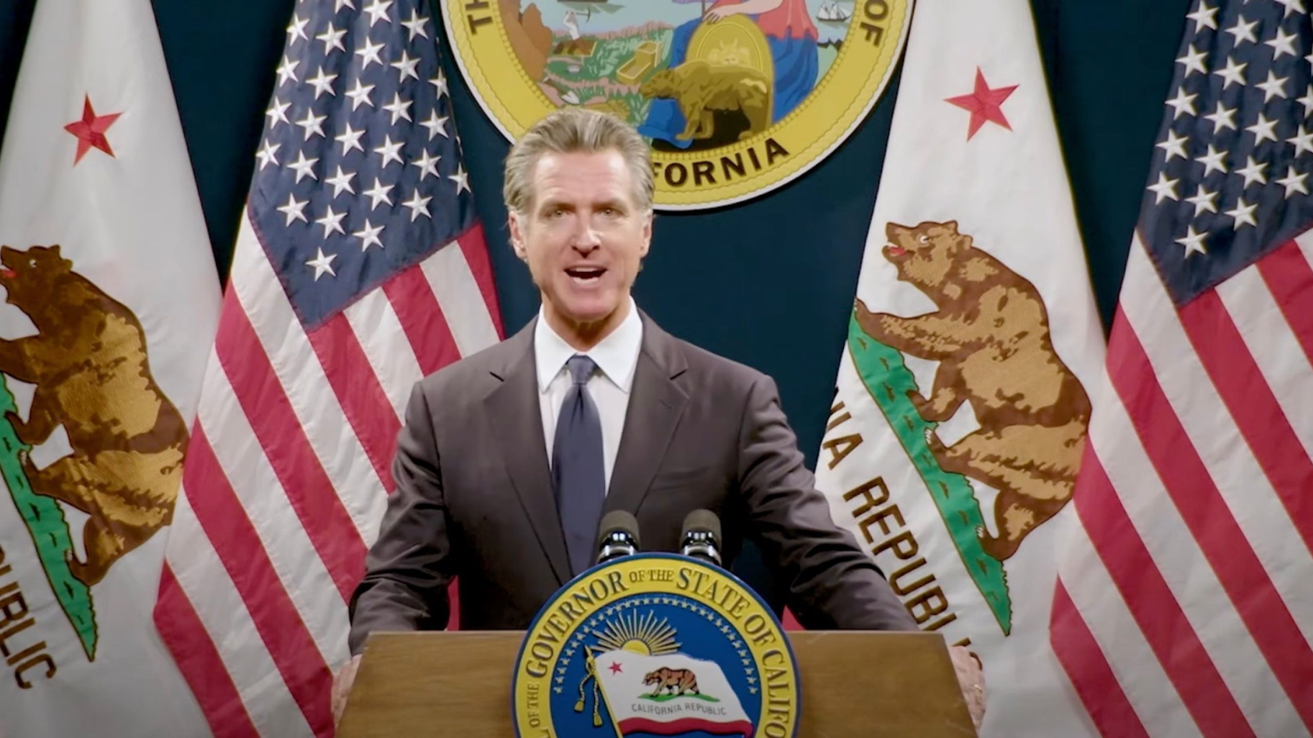 Gavin Newsom standing at a podium with the seal of the Governor of California, flanked by U.S. and California flags.