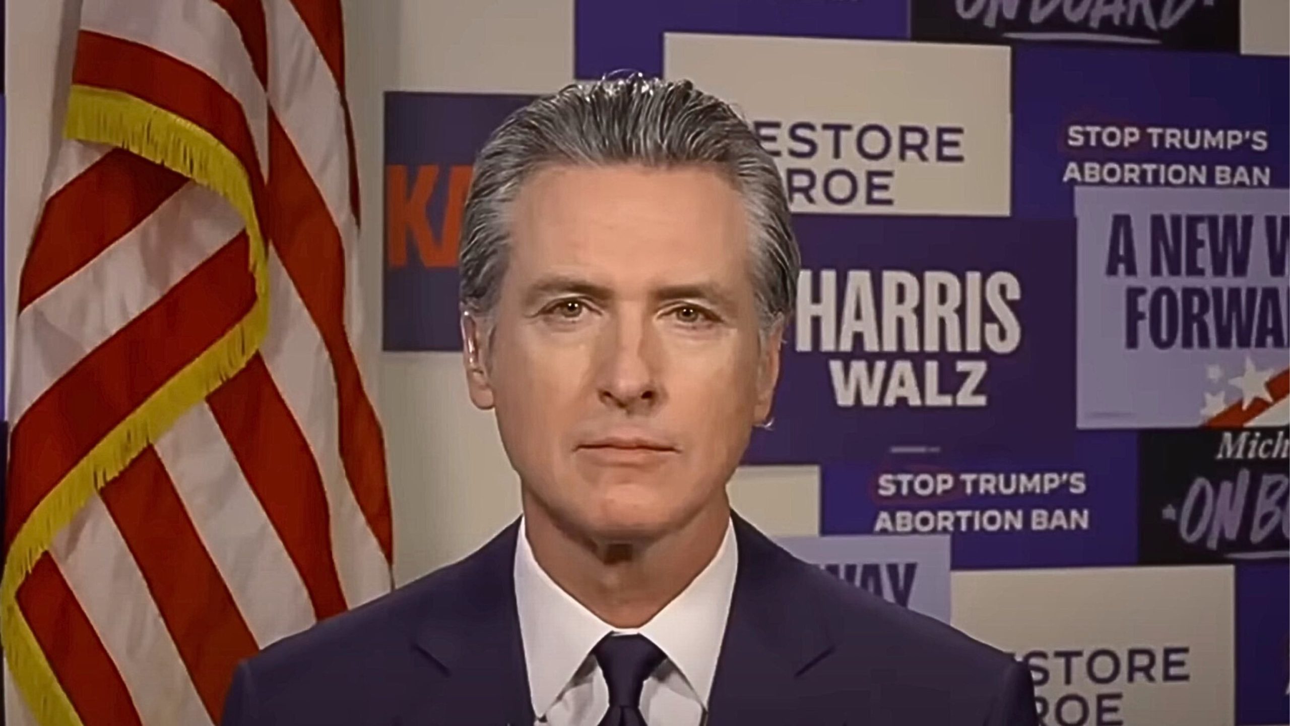 Gavin Newsom standing in front of an American flag and campaign posters with messages about restoring Roe and opposing Trump's abortion ban.