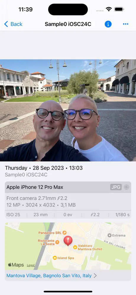 A selfie of two people smiling outdoors in a shopping area with Mediterranean-style architecture, captured on an Apple iPhone 12 Pro Max.