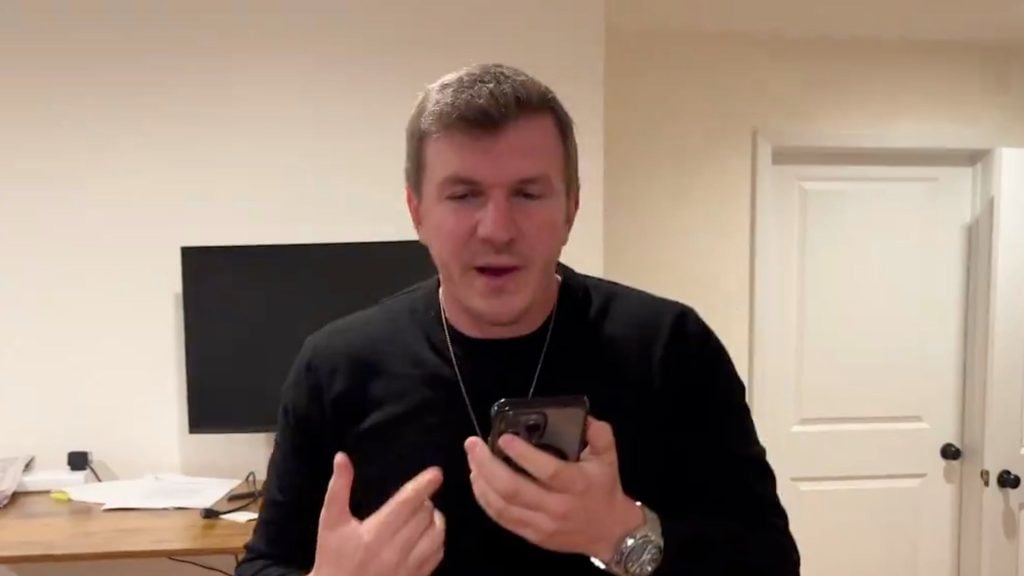 James O'Keefe holding a smartphone, standing in a room with a desk and a monitor in the background.