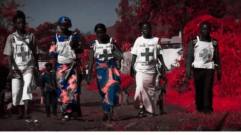 A group of people wearing vests with Red Cross symbols walking in an outdoor setting, with red foliage in the background.