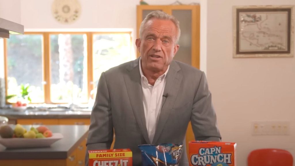 RFK Jr. in a suit stands in a kitchen with boxes of Cheez-It, Doritos, and Cap'n Crunch in front of him.