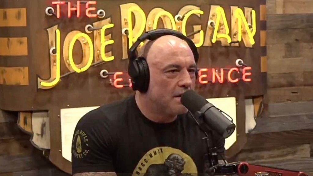 Joe Rogan wearing headphones speaks into a microphone in front of a sign that says "The Joe Rogan Experience."