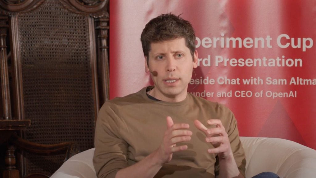 Sam Altman speaking into a microphone, seated indoors with a red backdrop featuring text.