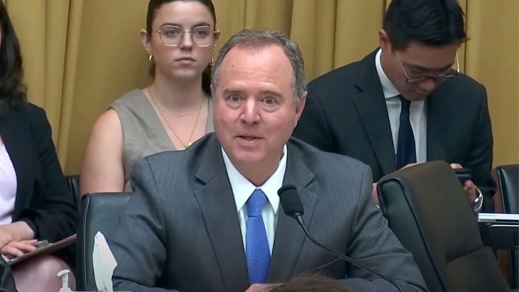 Adam Schiff speaks into a microphone at a formal event, with three individuals seated behind him, two of whom are using electronic devices.