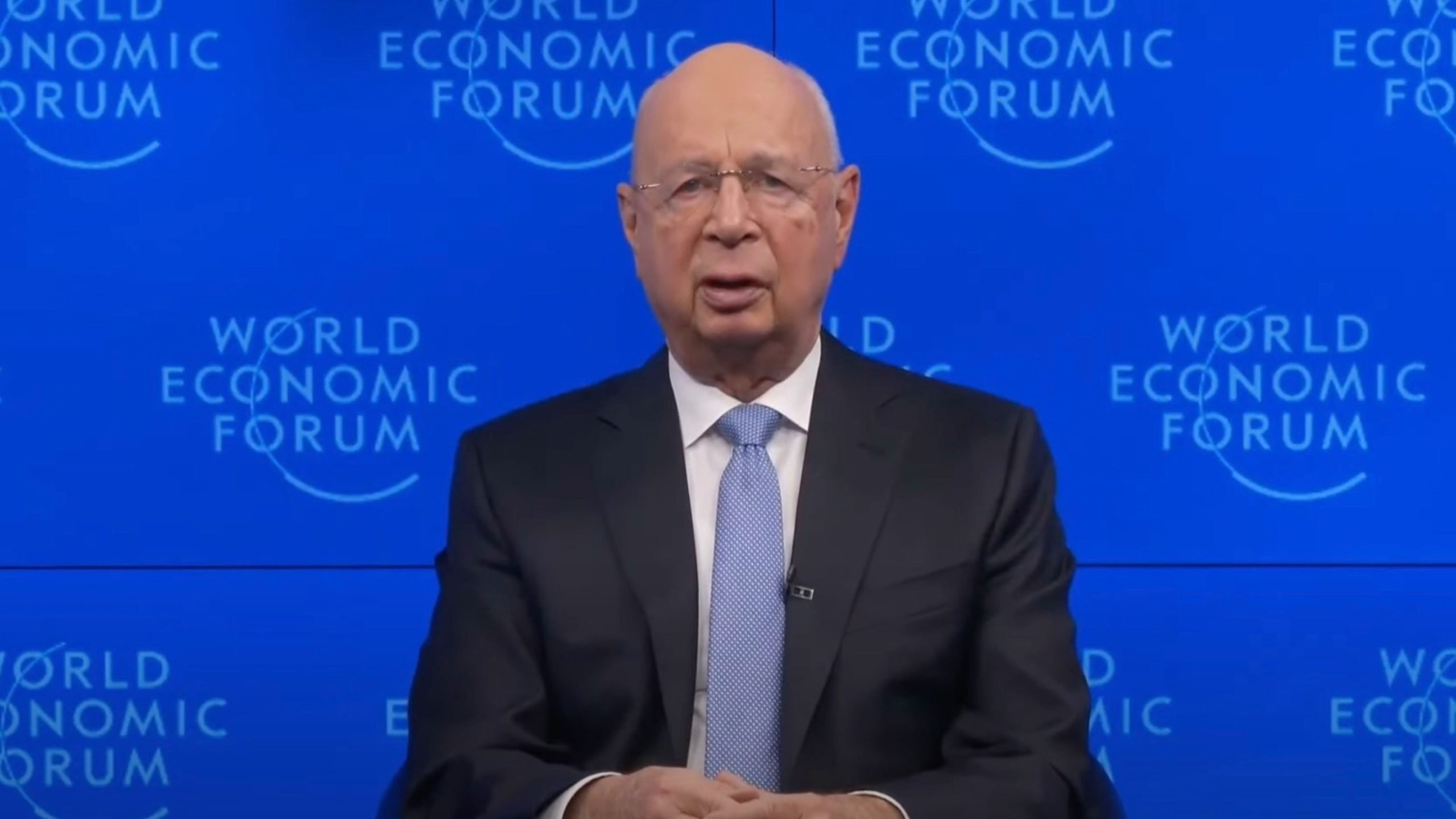 Klaus Schwab with a blue background featuring "World Economic Forum" logos.