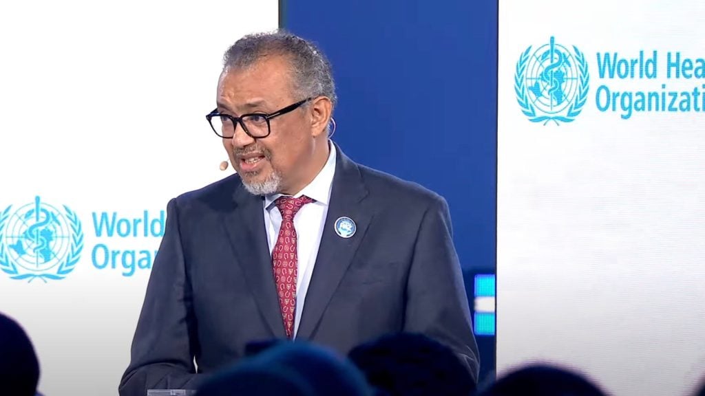 Tedros in a suit and red tie speaks at a podium with a badge on his jacket, in front of a screen displaying the World Health Organization logo.