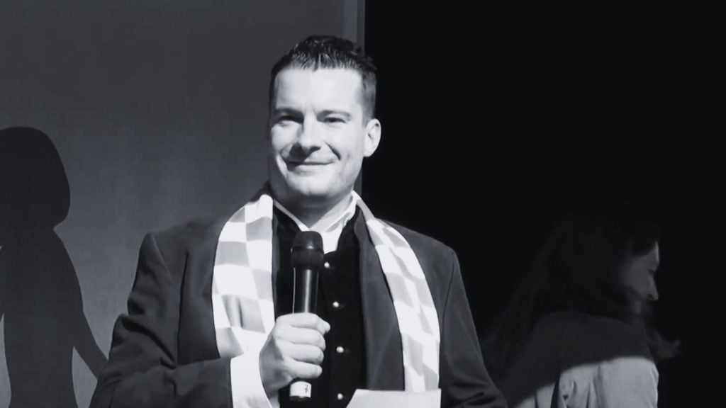 David Bendels holding a microphone, wearing a checkered scarf, and standing on a stage.