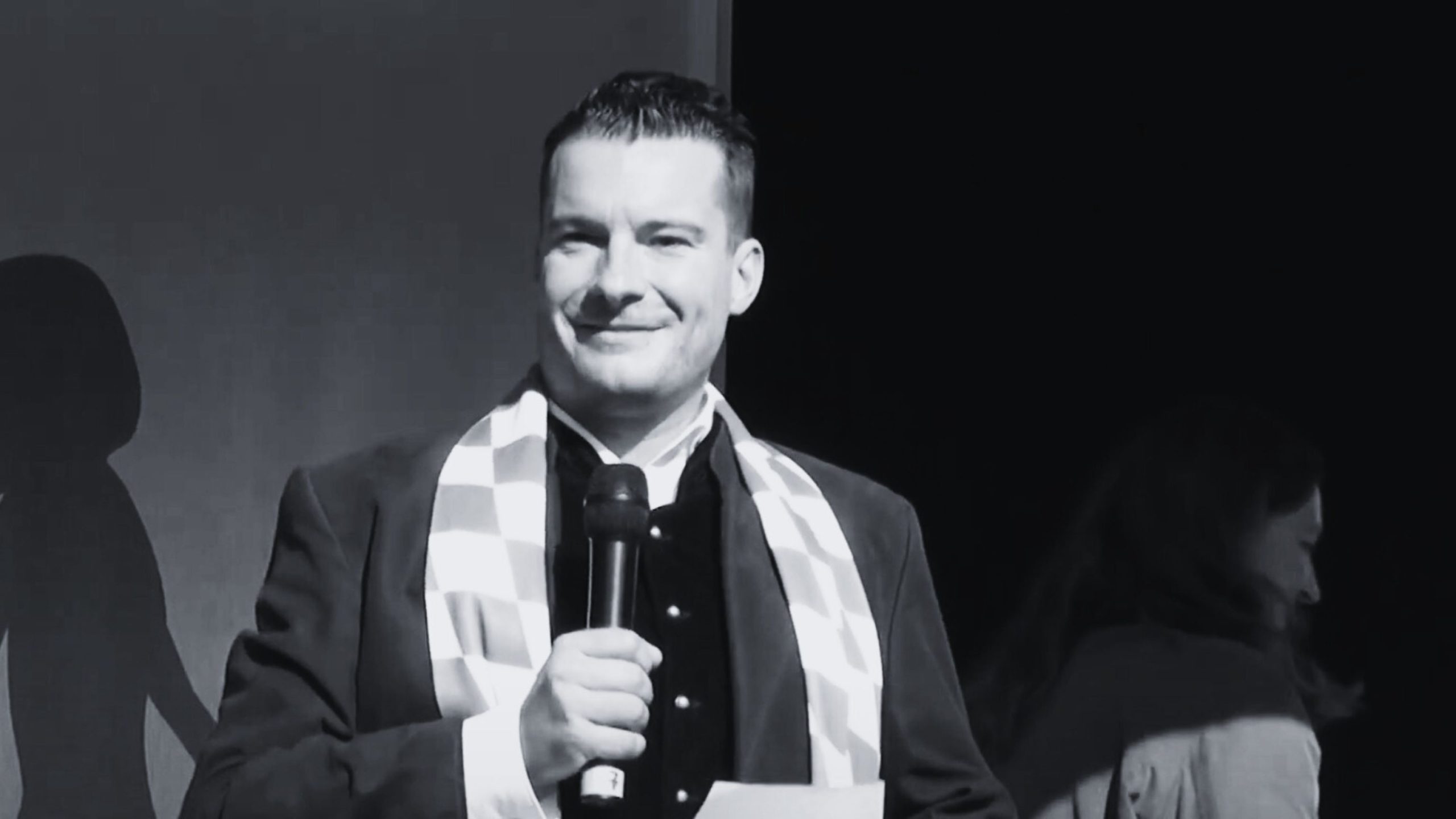 David Bendels holding a microphone, wearing a checkered scarf, and standing on a stage.