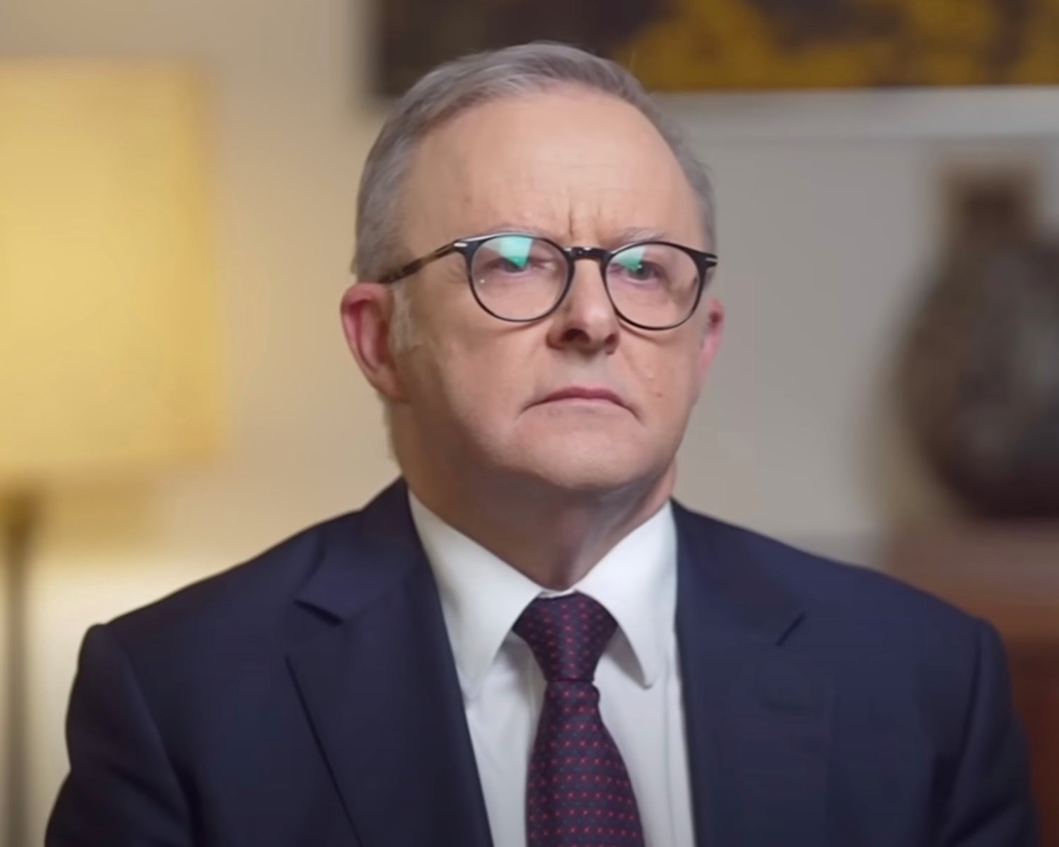 A man wearing glasses and a suit sitting indoors with a serious expression.