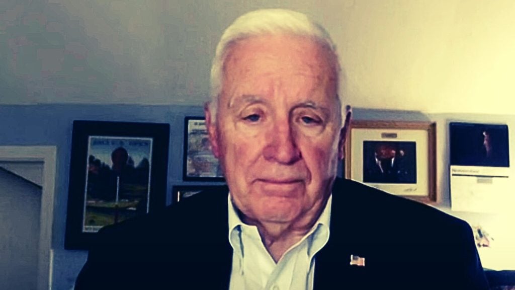 Tom Corbett with white hair wearing a dark suit and white shirt, standing in front of framed pictures on a wall.